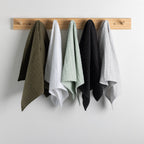 Four textured cotton tea towels in olive green, white, mint green, and black, hanging from a light wood peg rack against a plain white wall.