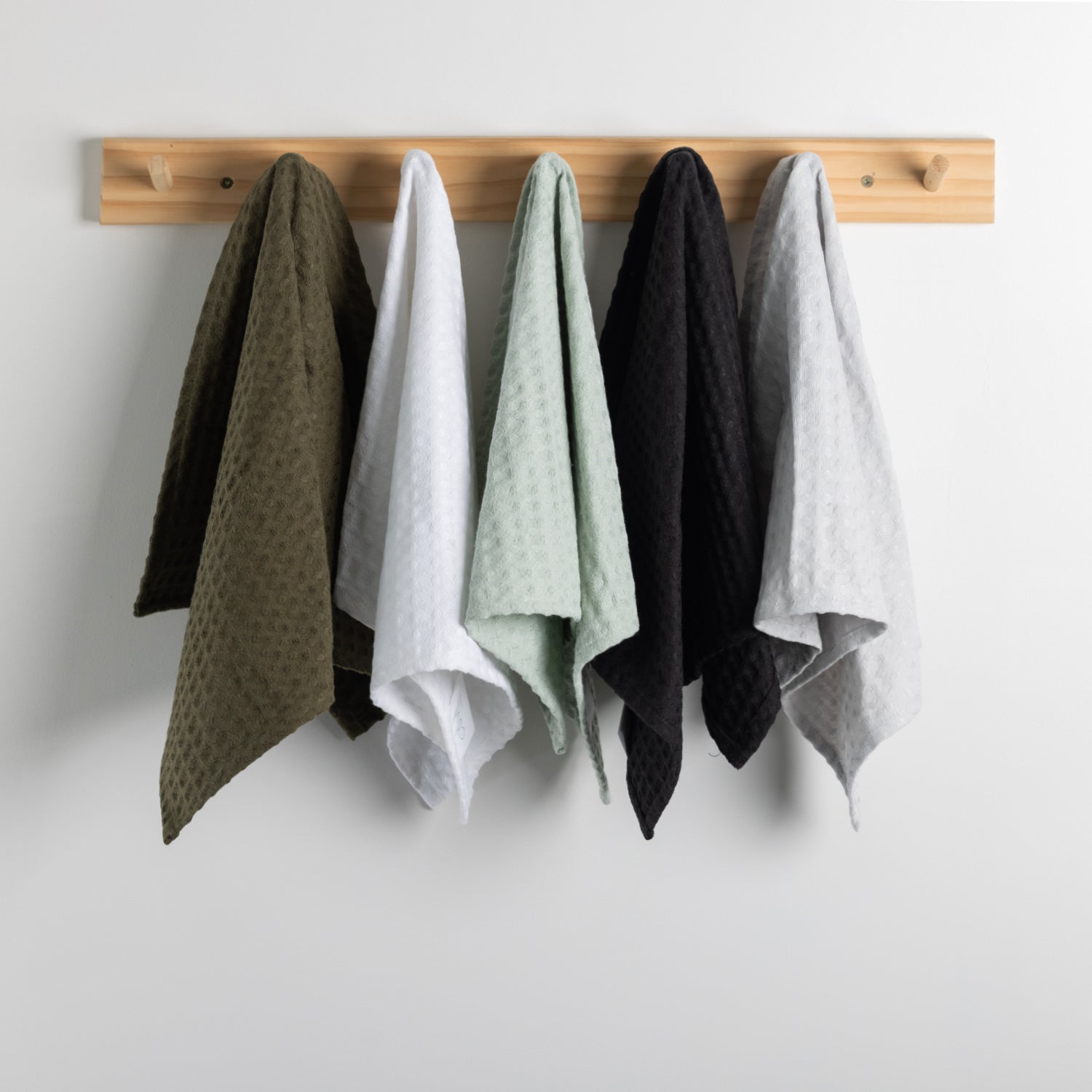 Four textured cotton tea towels in olive green, white, mint green, and black, hanging from a light wood peg rack against a plain white wall.
