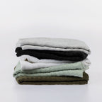 A stack of five folded textured cotton tea towels in light gray, black, white, sage green, and olive green, arranged neatly on a seamless white background.