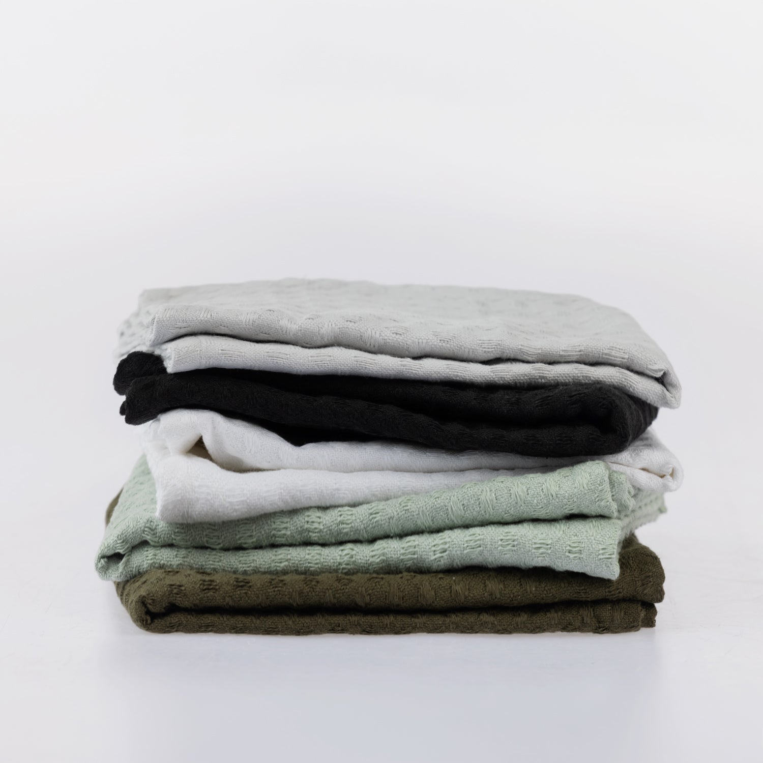 A stack of five folded textured cotton tea towels in light gray, black, white, sage green, and olive green, arranged neatly on a seamless white background.