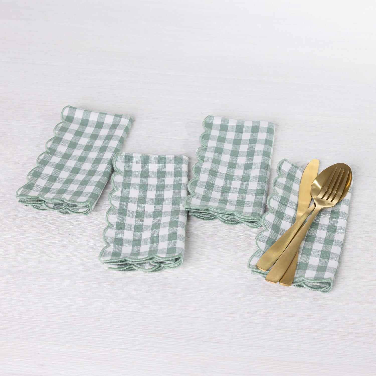 Gather Scallop Vichy Check napkin 4 pack in Green/Ivory arranged on a white table, with one napkin styled with gold-toned cutlery.