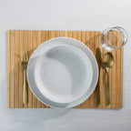 Overhead view of the Gather Bamboo Placemat in Natural, styled with white ceramic dinnerware, gold cutlery and a clear drinking glass.
