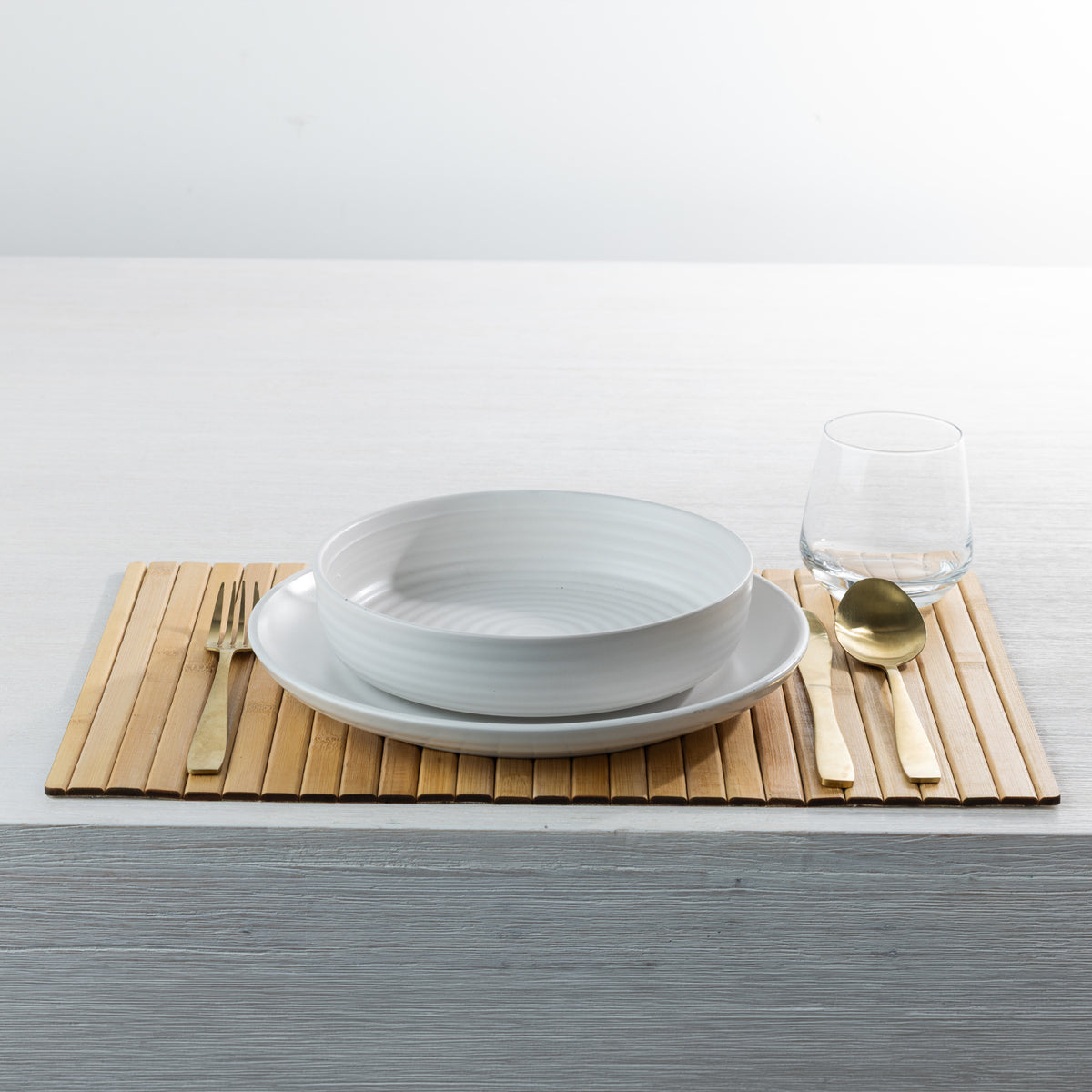 Gather Bamboo Placemat in Natural, styled on a white dining table with a white ceramic bowl and plate, gold cutlery and a clear drinking glass.