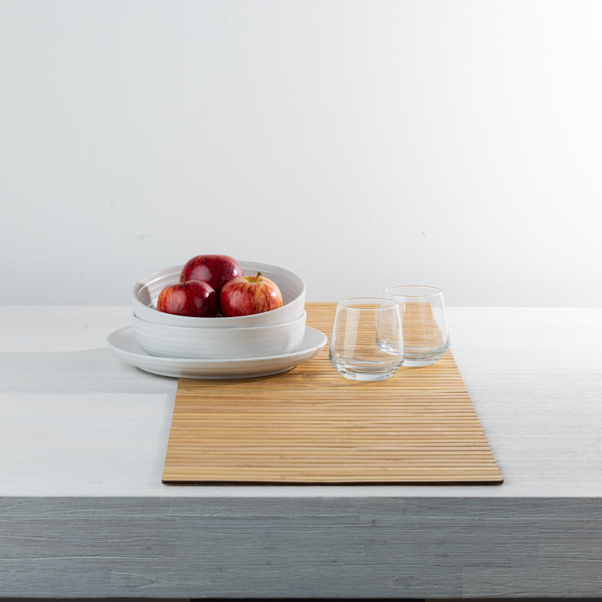 Gather Bamboo Runner in Natural, styled on a white dining table with white ceramic bowls filled with red apples and two clear drinking glasses, showcasing its warm bamboo finish.