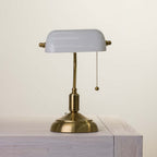 A classic brass desk lamp with a white glass shade, resting on a light-colored wooden surface against a neutral cream background.