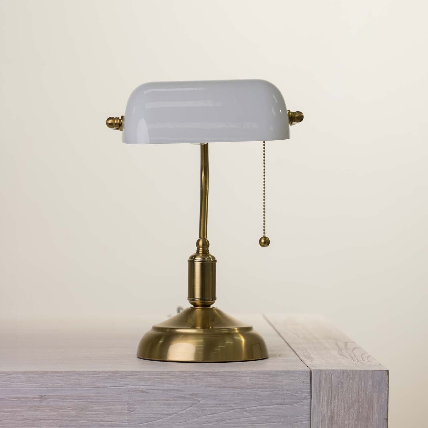 A classic brass desk lamp with a white glass shade, resting on a light-colored wooden surface against a neutral cream background.