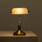 A classic brass table lamp with a glowing white glass shade, resting on a light wood surface against a dark background.