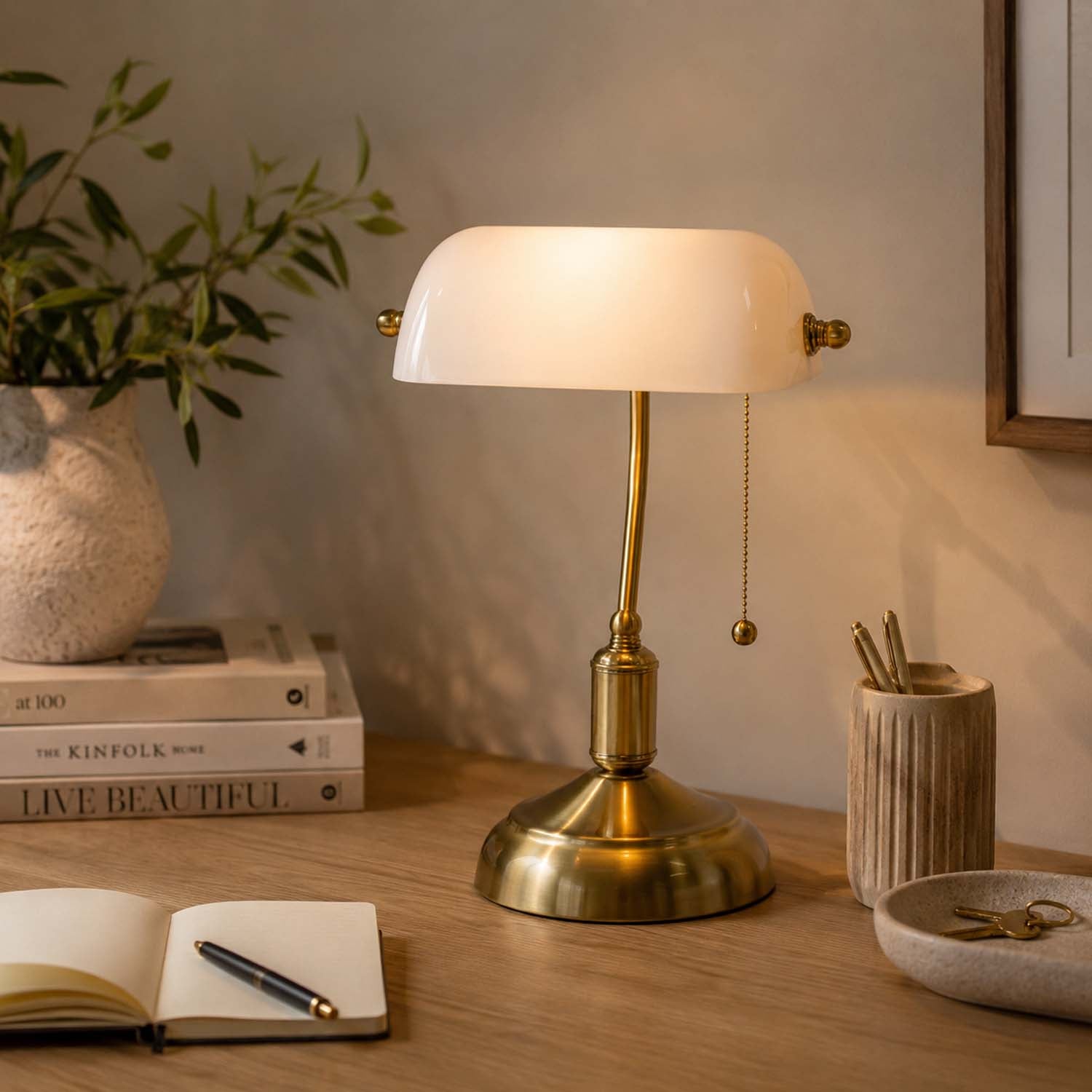 A gold table lamp with a white glass dome shade sits on a wooden desk in a warm, styled room setting with books, a plant, and stationery.