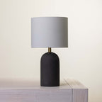 A modern table lamp with a textured black cylindrical base and a fabric drum shade, resting on a light gray wooden surface against a warm off-white background.