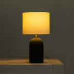 A table lamp with a glowing warm yellow fabric drum shade and a dark textured cylindrical base, shown sitting on a light wood surface against a dark gray wall.