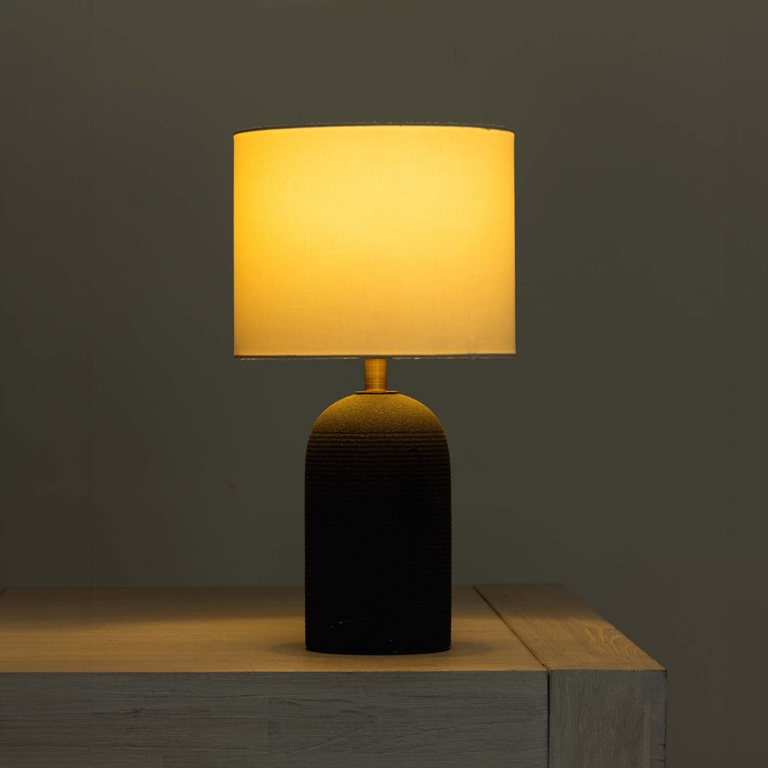 A table lamp with a glowing warm yellow fabric drum shade and a dark textured cylindrical base, shown sitting on a light wood surface against a dark gray wall.