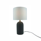 A modern table lamp with a textured dark cylindrical base, a gold-finished neck, and a white drum shade, shown on a plain white background with its power cord visible.
