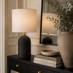 A table lamp with a textured dark base and a warm off-white fabric drum shade sits on a dark wood console table in a styled interior. A matching lamp is reflected in a large black-framed mirror, alongside books, a small bowl, and a large ceramic vase with greenery.