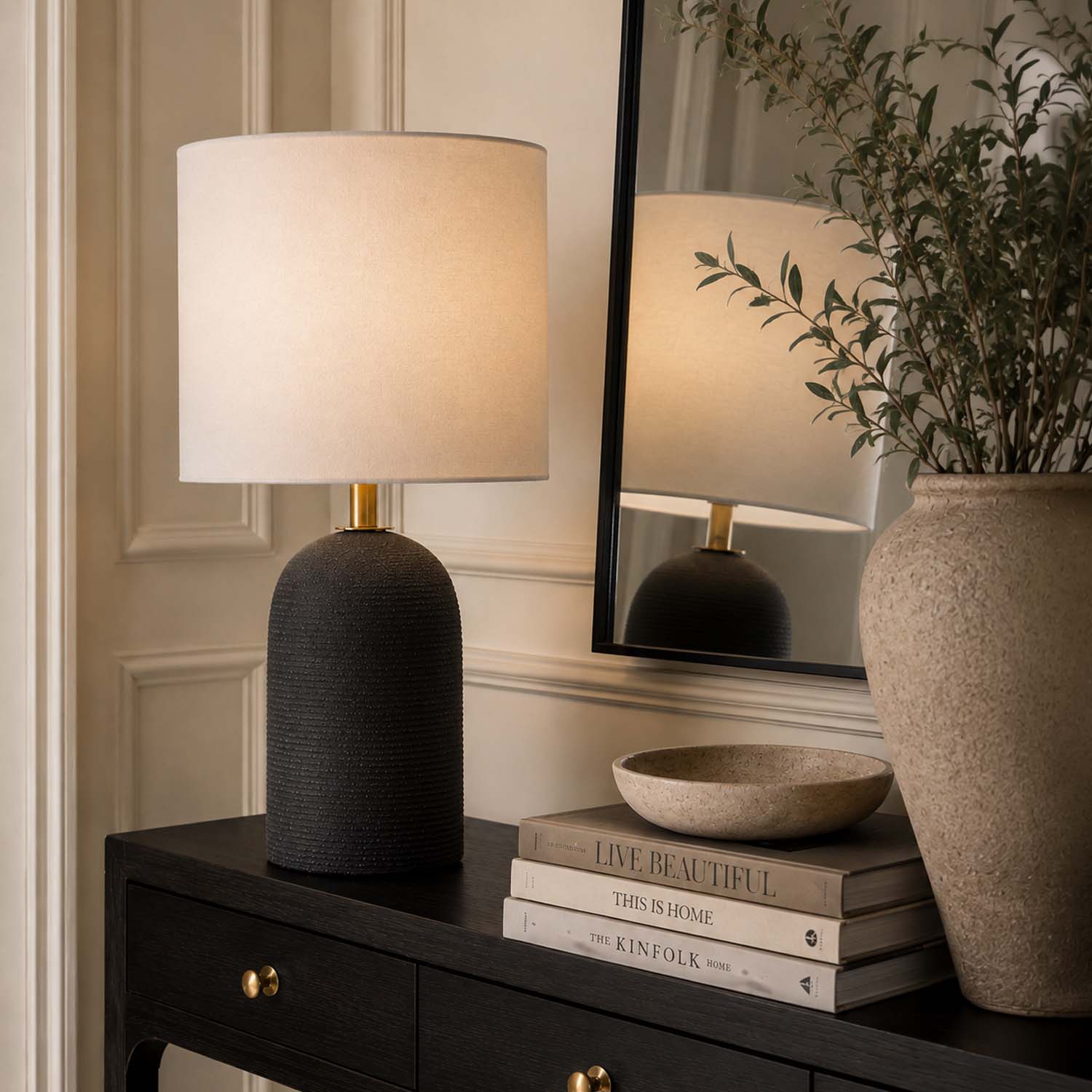 A table lamp with a textured dark base and a warm off-white fabric drum shade sits on a dark wood console table in a styled interior. A matching lamp is reflected in a large black-framed mirror, alongside books, a small bowl, and a large ceramic vase with greenery.