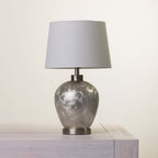 A table lamp with a textured light fabric drum shade and a metallic silver-gray marbled ceramic base, shown sitting on a light-colored wooden surface against a plain off-white background.