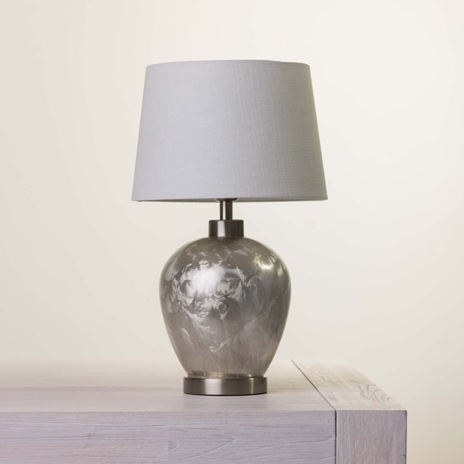 A table lamp with a textured light fabric drum shade and a metallic silver-gray marbled ceramic base, shown sitting on a light-colored wooden surface against a plain off-white background.
