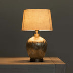 A table lamp with a textured linen drum shade and a metallic ceramic base, illuminated against a dark gray background.