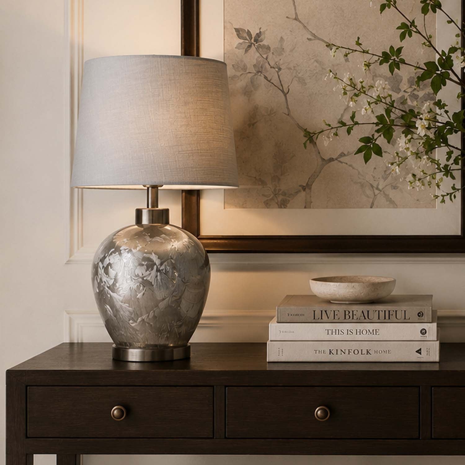A table lamp with a textured metallic silver ceramic base and a light beige linen drum shade, styled on a dark wood console table next to a stack of books and a small bowl, with a framed botanical print in the background.