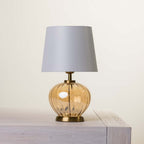 A table lamp with a light fabric drum shade, a ribbed amber glass base, and a brass metal collar and base, shown resting on a light gray wooden surface against a warm off-white background.
