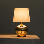 A table lamp with a warm glowing rectangular fabric shade and a reflective amber glass base, shown lit on a wooden surface against a dark gray background.