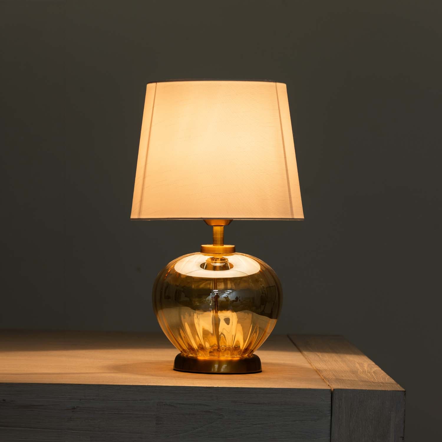 A table lamp with a warm glowing rectangular fabric shade and a reflective amber glass base, shown lit on a wooden surface against a dark gray background.