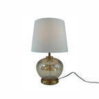 A table lamp with a white fabric drum shade, a clear amber glass bulbous base, and brass-toned metal hardware, shown on a plain white background with a black power cord extending to the right.