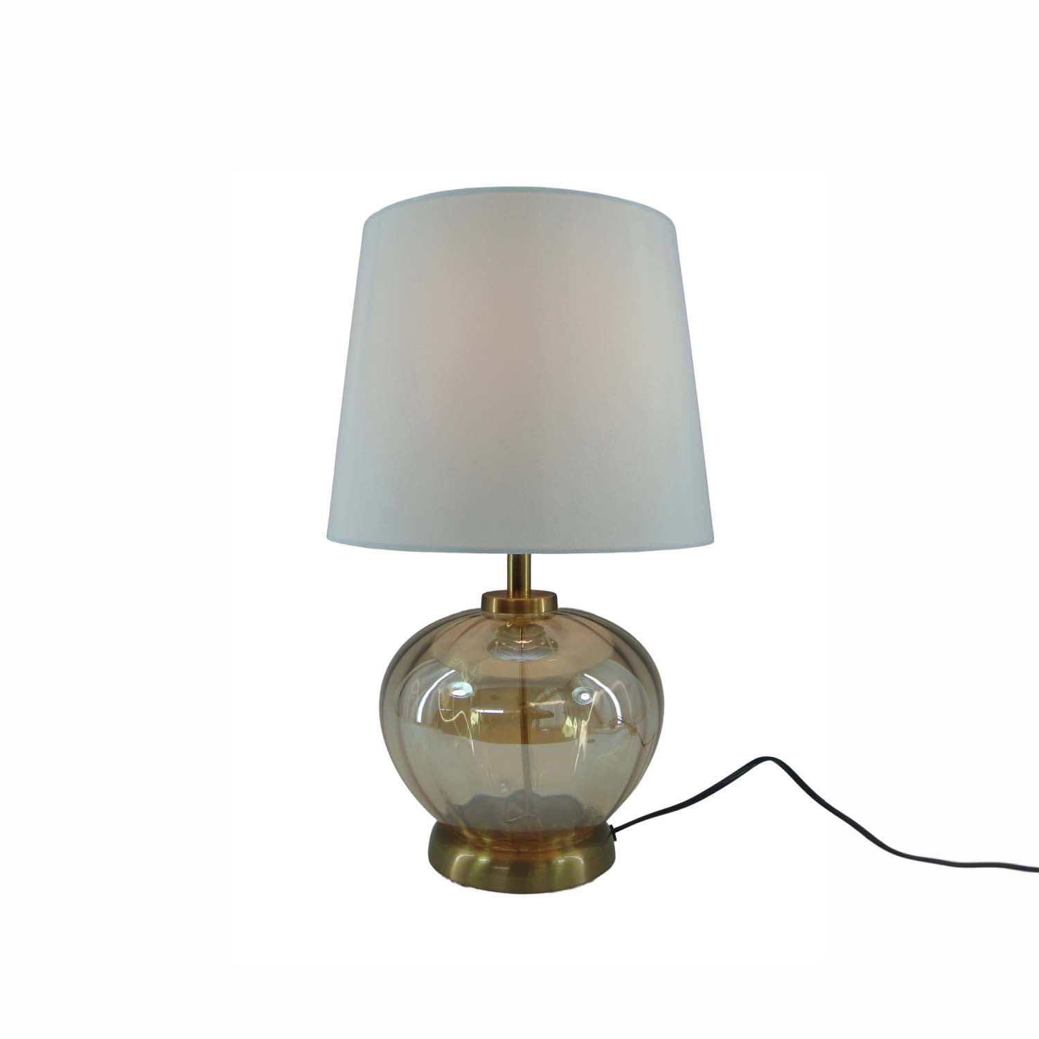 A table lamp with a white fabric drum shade, a clear amber glass bulbous base, and brass-toned metal hardware, shown on a plain white background with a black power cord extending to the right.