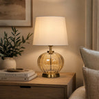 A table lamp with a warm amber glass ribbed base and a cream fabric shade, illuminated on a wooden nightstand in a cozy room setting.