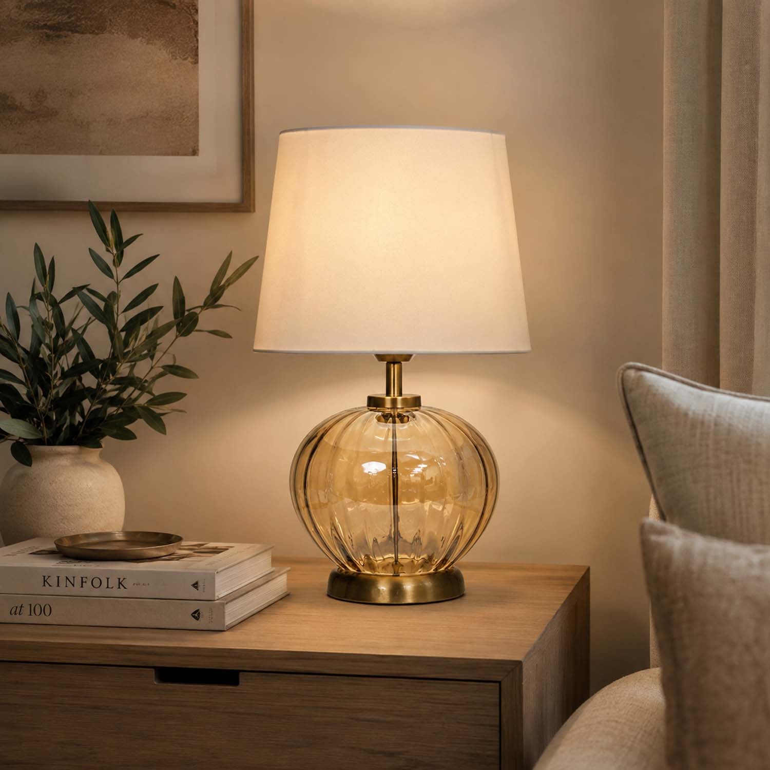 A table lamp with a warm amber glass ribbed base and a cream fabric shade, illuminated on a wooden nightstand in a cozy room setting.
