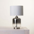 A table lamp with a gray gradient glass base and a fabric drum shade, displayed on a light wood surface against a warm off-white background.