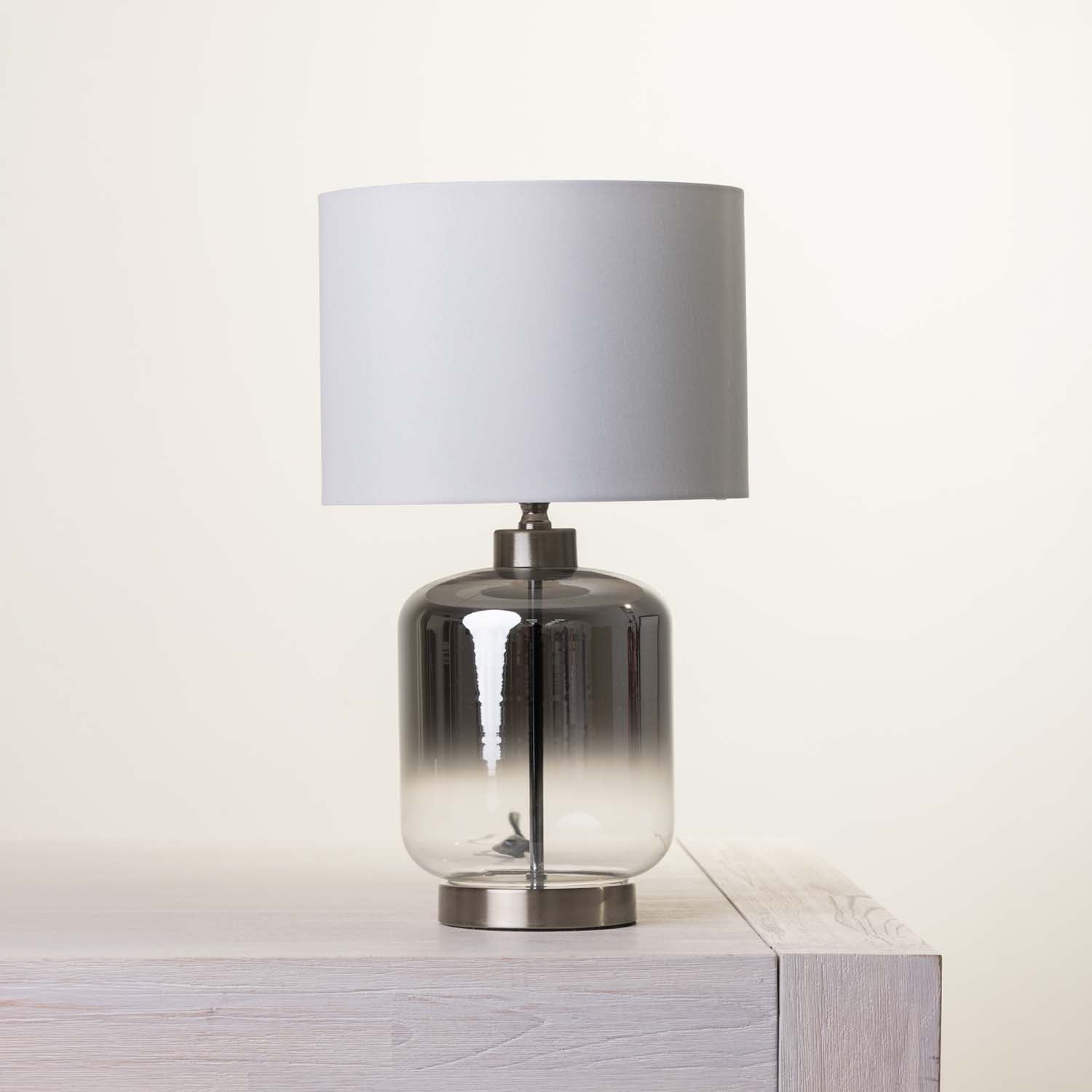 A table lamp with a gray gradient glass base and a fabric drum shade, displayed on a light wood surface against a warm off-white background.