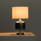 A modern table lamp with a glowing warm white fabric drum shade, a glossy grey-to-clear glass cylindrical base, and a metal collar and base, shown resting on a light wood surface against a dark gray wall.