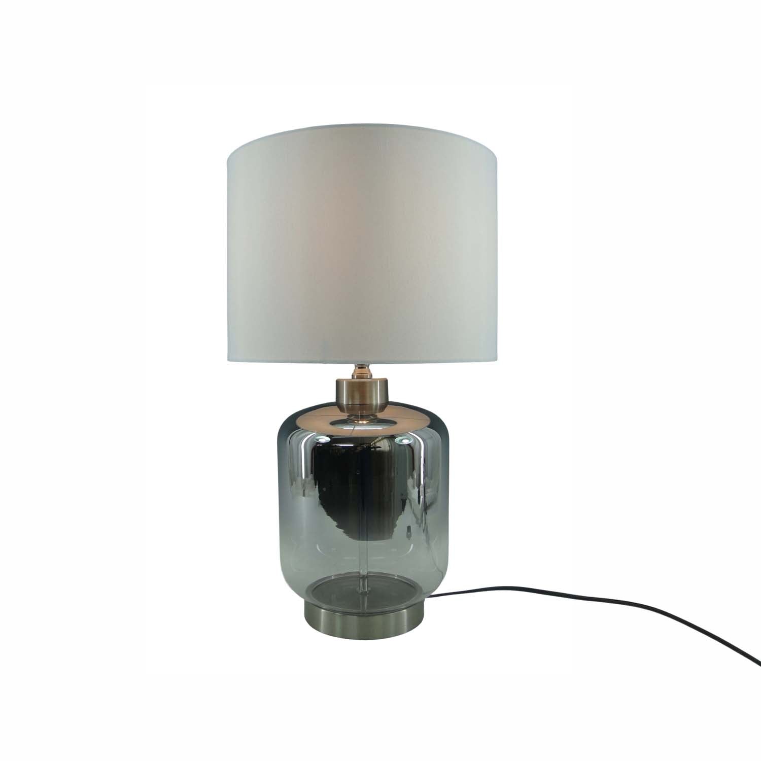 A modern table lamp featuring a cylindrical smoky gray glass base, a brushed silver metal collar, and a white fabric drum shade, shown against a plain white background with its power cord visible.