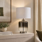 A table lamp with a white drum shade and a smoky gradient glass base on a light wood console table in a warm, neutral-toned room.