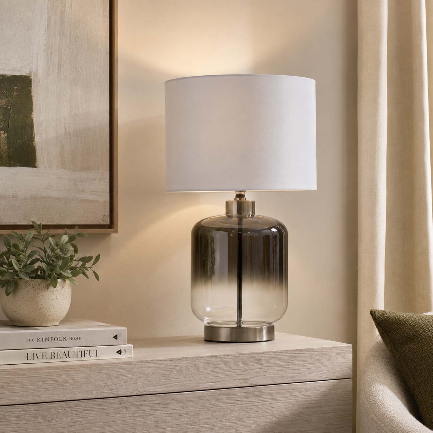 A table lamp with a white drum shade and a smoky gradient glass base on a light wood console table in a warm, neutral-toned room.