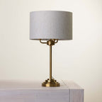 A table lamp with a cylindrical light fabric shade and a polished brass-finished metal base, shown resting on a light gray wooden surface against a plain off-white background.