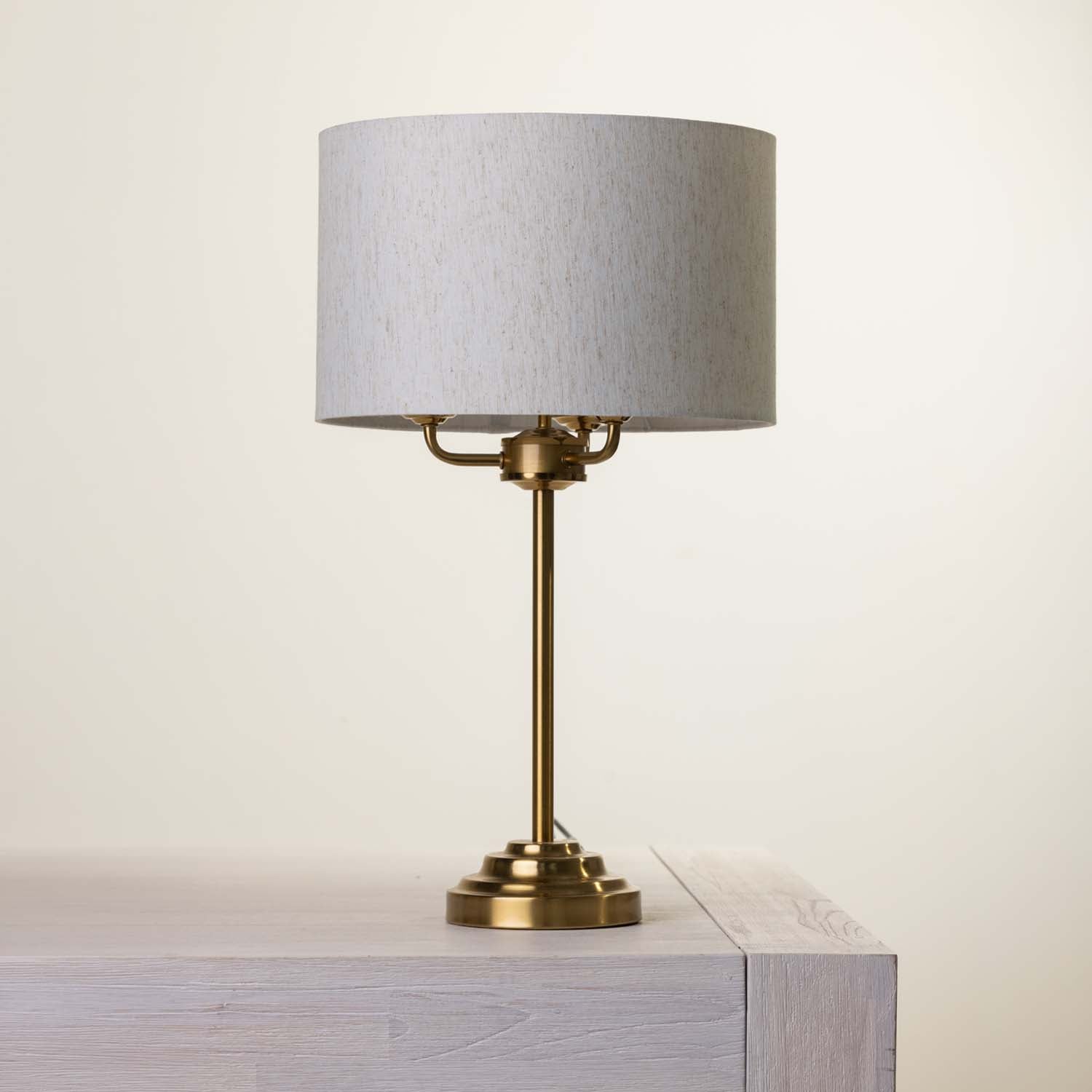 A table lamp with a cylindrical light fabric shade and a polished brass-finished metal base, shown resting on a light gray wooden surface against a plain off-white background.