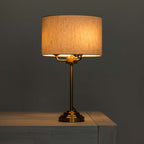 A table lamp with a warm glowing drum shade, a brass-finished metal stem and base, shown lit on a wooden surface against a dark gray background.