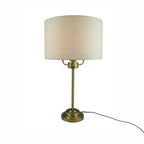 A table lamp with a cylindrical off-white fabric shade and a brass-finished metal base and stem, shown on a plain white background with its power cord visible.