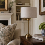A brass table lamp with a textured light beige drum shade sits on a dark wood side table in a cozy, classic living room setting next to a floral throw pillow.