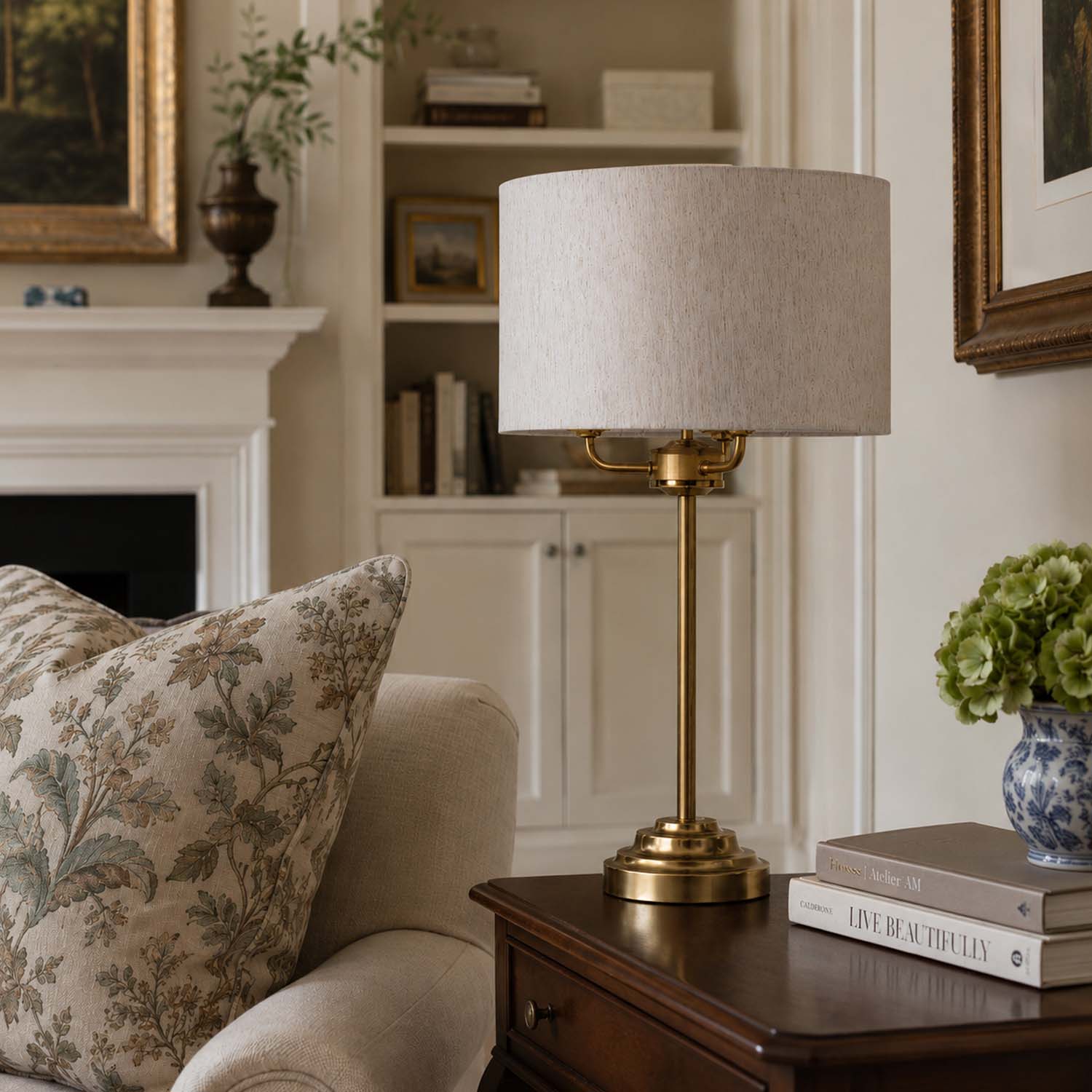 A brass table lamp with a textured light beige drum shade sits on a dark wood side table in a cozy, classic living room setting next to a floral throw pillow.