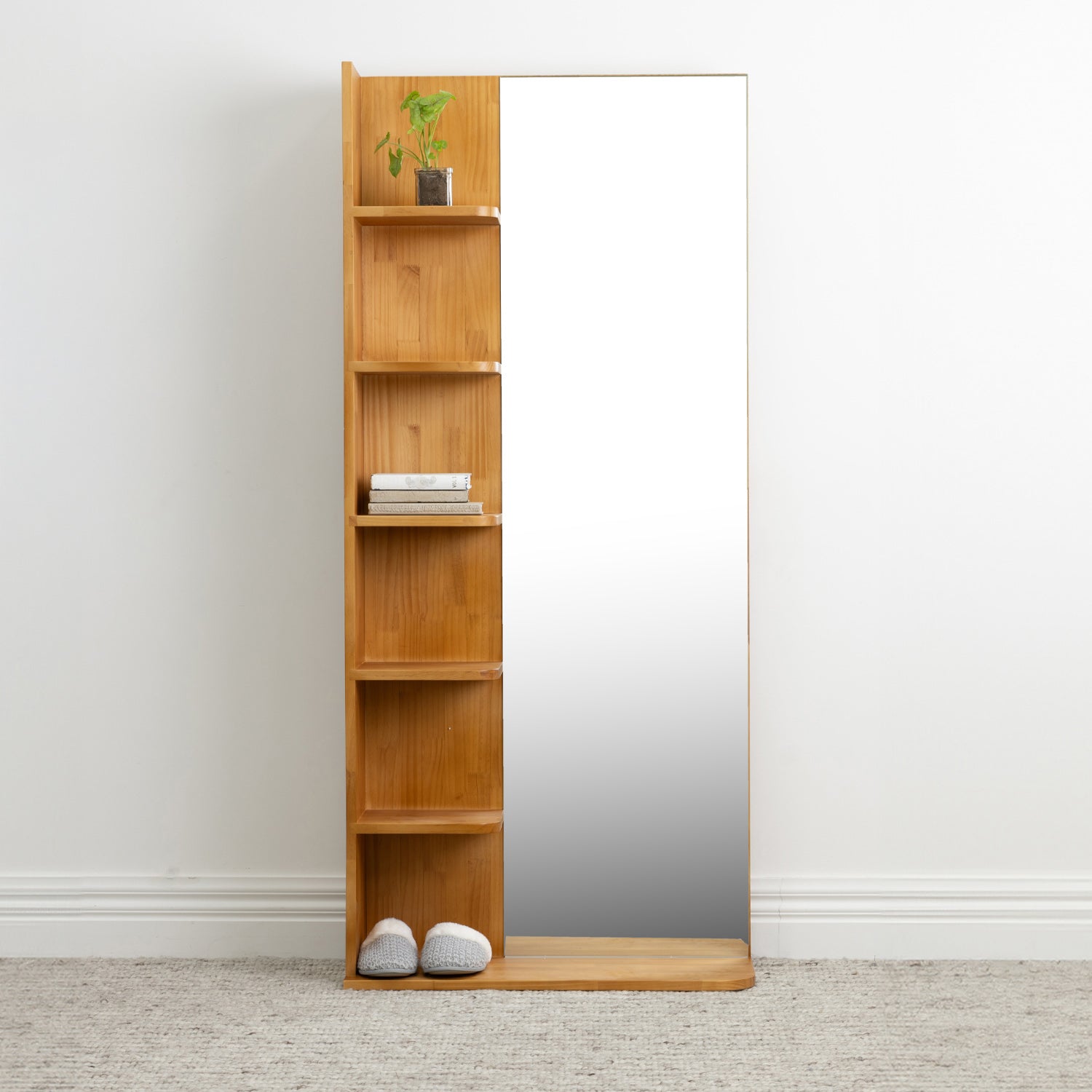 A tall wooden floor mirror with five open shelves on the left side, styled with a small potted plant, books, and a pair of slippers on a light gray carpet against a white wall.