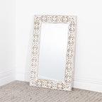 A tall rectangular wall mirror with an ornate carved white frame, leaning against a white wall on a textured beige carpet.