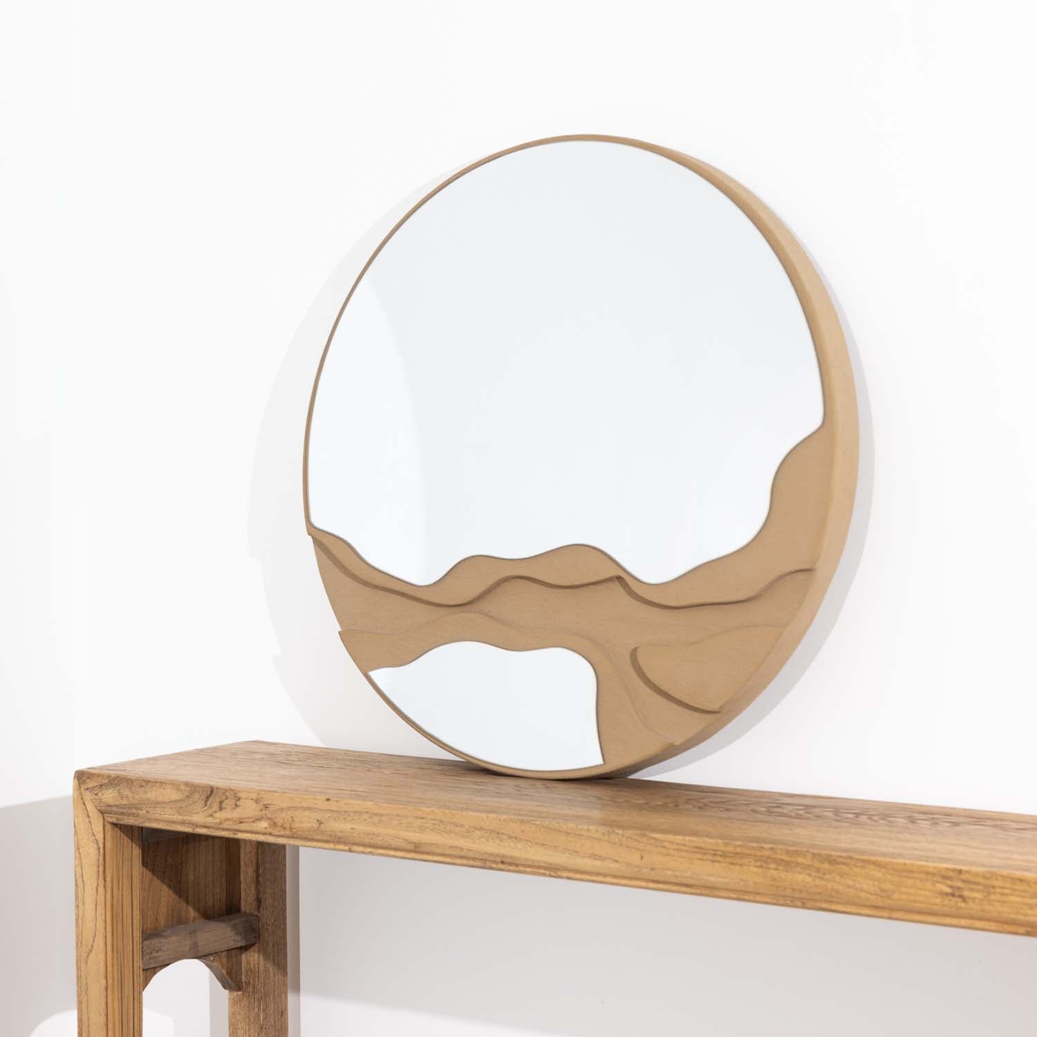 Round decorative mirror with an organic sculpted natural-toned frame and abstract layered detailing, styled on a wooden console table against a white wall.