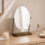 Gold tabletop vanity mirror with an oval frameless design displayed on a wooden vanity alongside candles, perfume bottles, and decorative accessories.