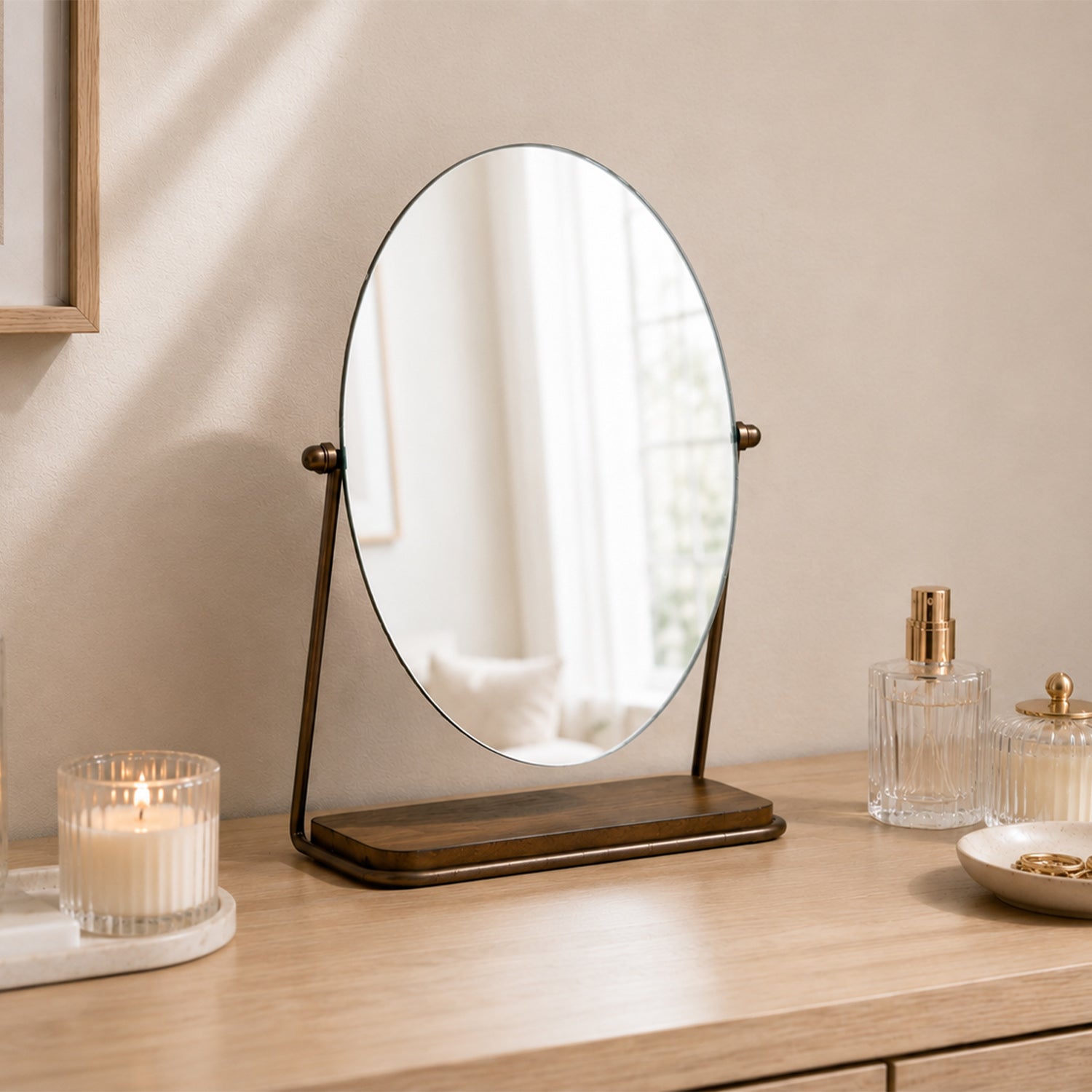 Gold tabletop vanity mirror with an oval frameless design displayed on a wooden vanity alongside candles, perfume bottles, and decorative accessories.