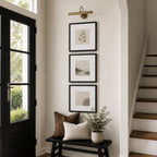 A bright, modern entryway features a black bench with decorative pillows and a potted plant beneath three vertically stacked black-framed artworks, accented by a brass picture light and a staircase nearby.