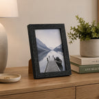 A black woodgrain photo frame displaying a scenic lake photograph is styled on a wooden console table beside a table lamp, stacked books, and a potted plant.