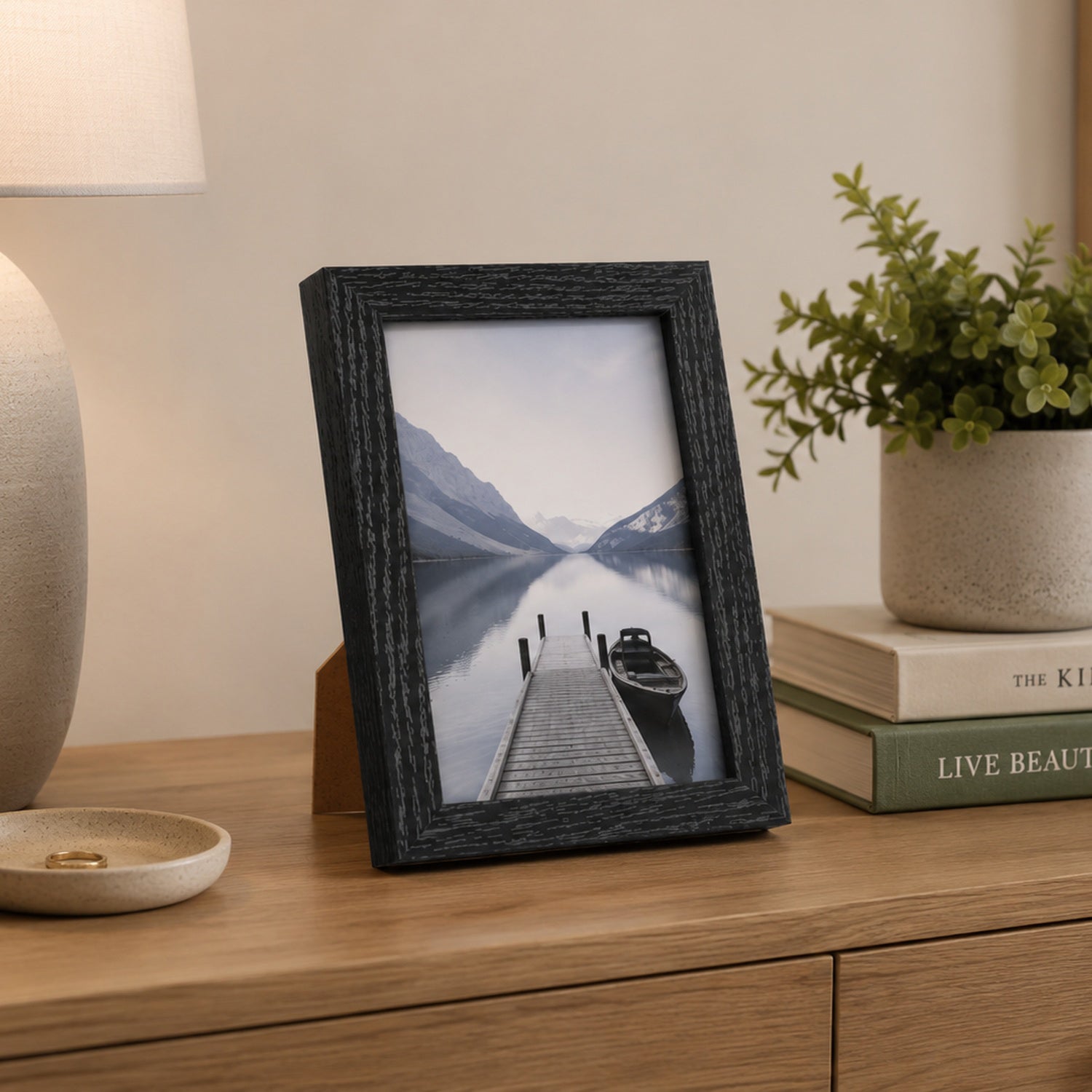 A black woodgrain photo frame displaying a scenic lake photograph is styled on a wooden console table beside a table lamp, stacked books, and a potted plant.