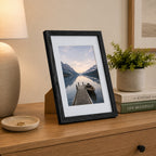A black woodgrain photo frame displaying a tranquil lakeside landscape is styled on a wooden console table beside a lamp, potted plant, and stacked books in a warm, contemporary interior.